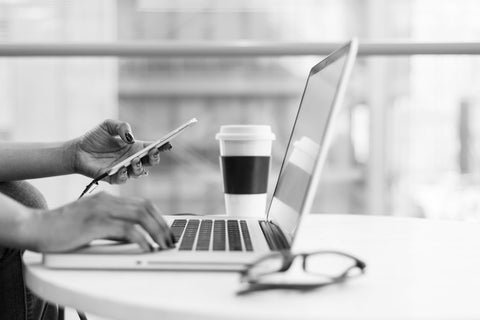 Black and white photo of Black woman typing on a laptop while checking her phone, with her glasses and a coffee on the white table.