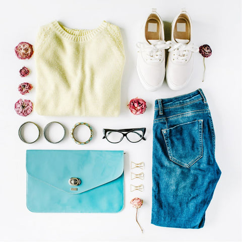 Flat lay of yellow sweater with blue jeans, white sneakers, jewelry, glasses, and an aqua colored clutch.