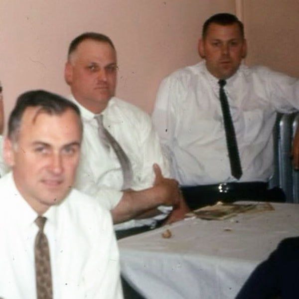 An old photo of three middle aged men in white shirts and skinny black ties sitting at a dining table