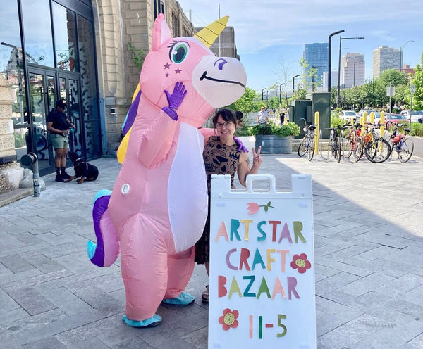 Megan at Cherry Street Pier with the Art Star Unicorn Mascot in front of a sign that says Art Star Craft Bazaar, 11-5.