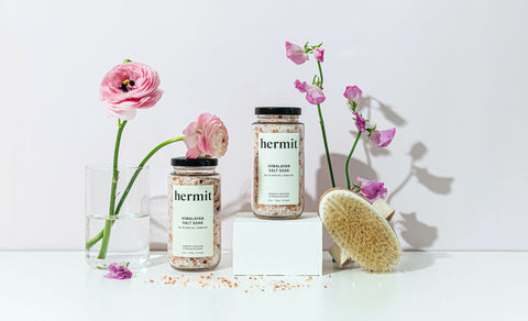 Hermit Goods products on display - two jars of the Himalayan Salt Soak and a Body Brush with two vases of flowers on white background