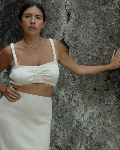 Tan female model in white skirt and white top with hand placed on a textured gray rock