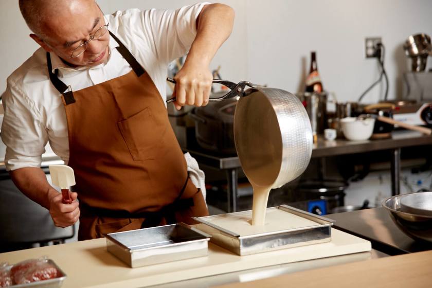 Man in brown waterproof apron pouring batter into dish in a kitchen