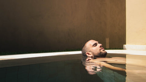 Dark haired, bearded man relaxing in a calm pool tub jacuzzi by himself, with just his head above the water. He is calm and relaxed.