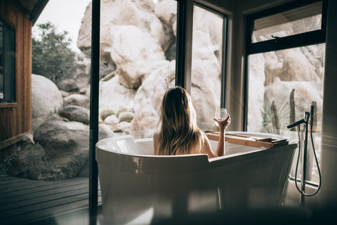 Woman in a white bathtub, holding a wine glass with her back facing us, looking outside towards a rock sculpture. Clean, minimalist bathroom aethetic.