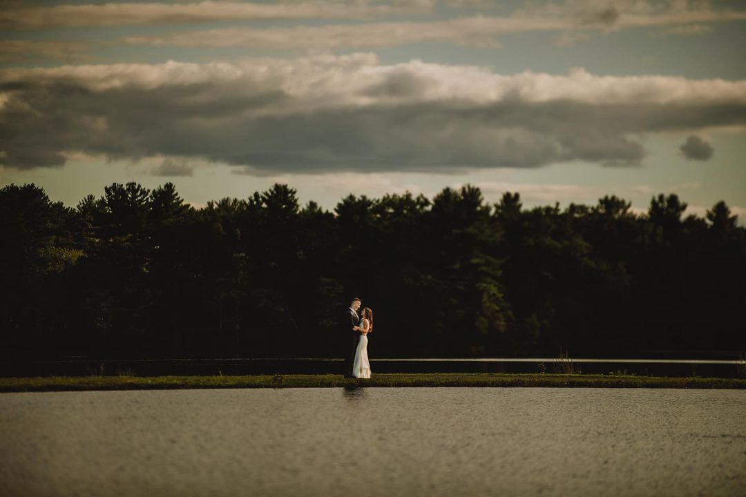 a couple standing infront of a forest beside a lake during the dusk 