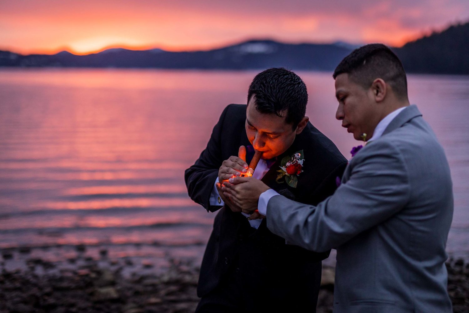 A groom with one of his groomsman lighting up a cigar with a waterfront background