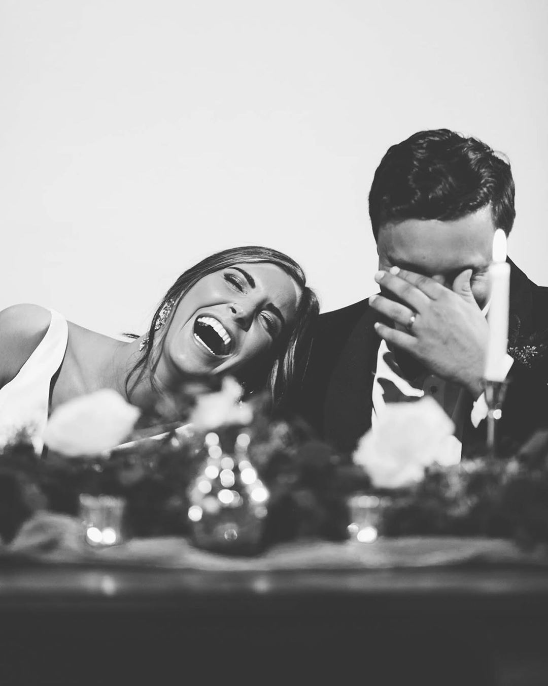 Black and white portrait of a couple laughing over the dinner table with the bride leaning over the groom’s shoulder