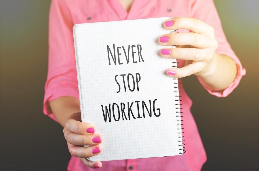 woman with pink nails and shirt holding a notebook that says never stop working