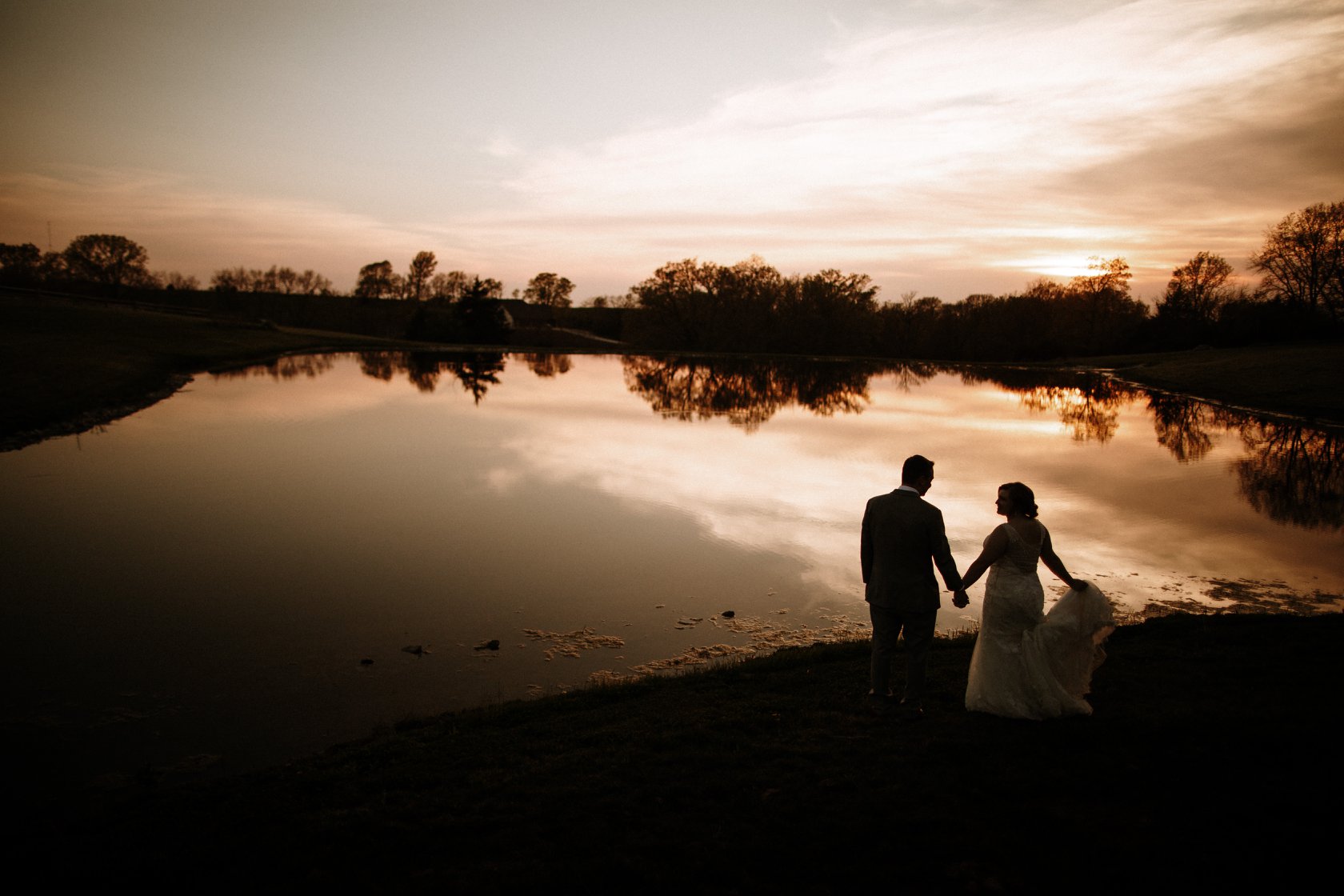 A bride and groom holding hands and posing in front of a lake during sunset