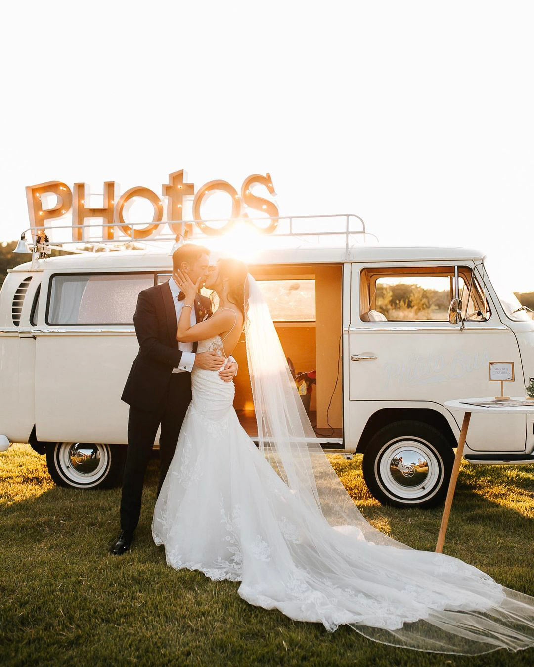 a couple standing in front of a van in their wedding attire while the sun shines from behind