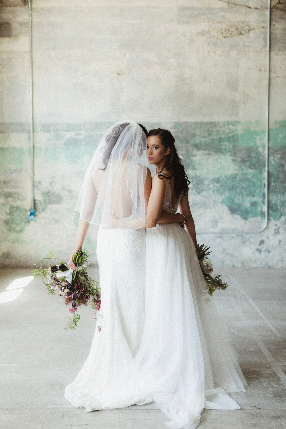 two brides in their wedding attire standing holding the wedding bouquets