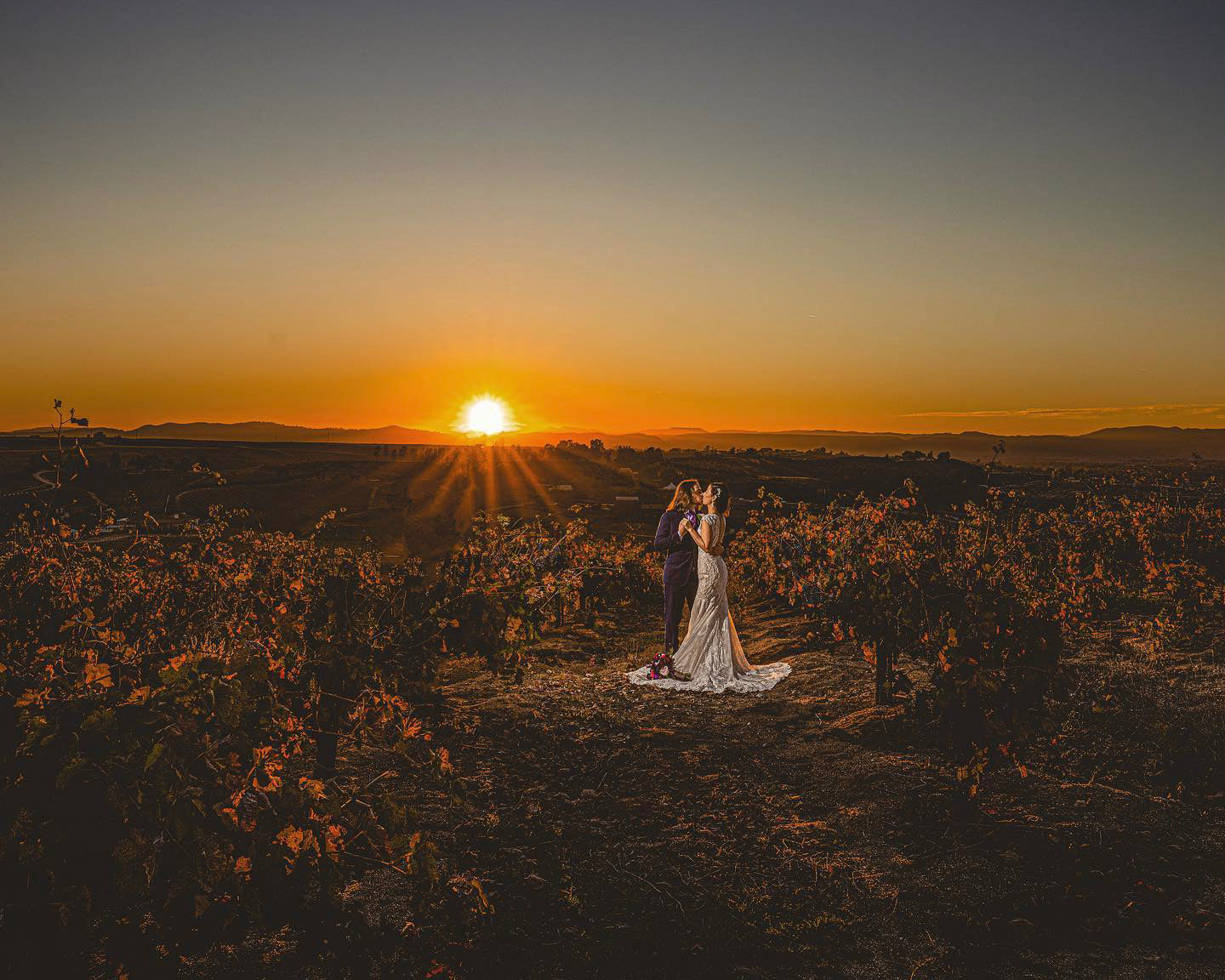 a newlywed couple kissing in the middle of a field during sunset