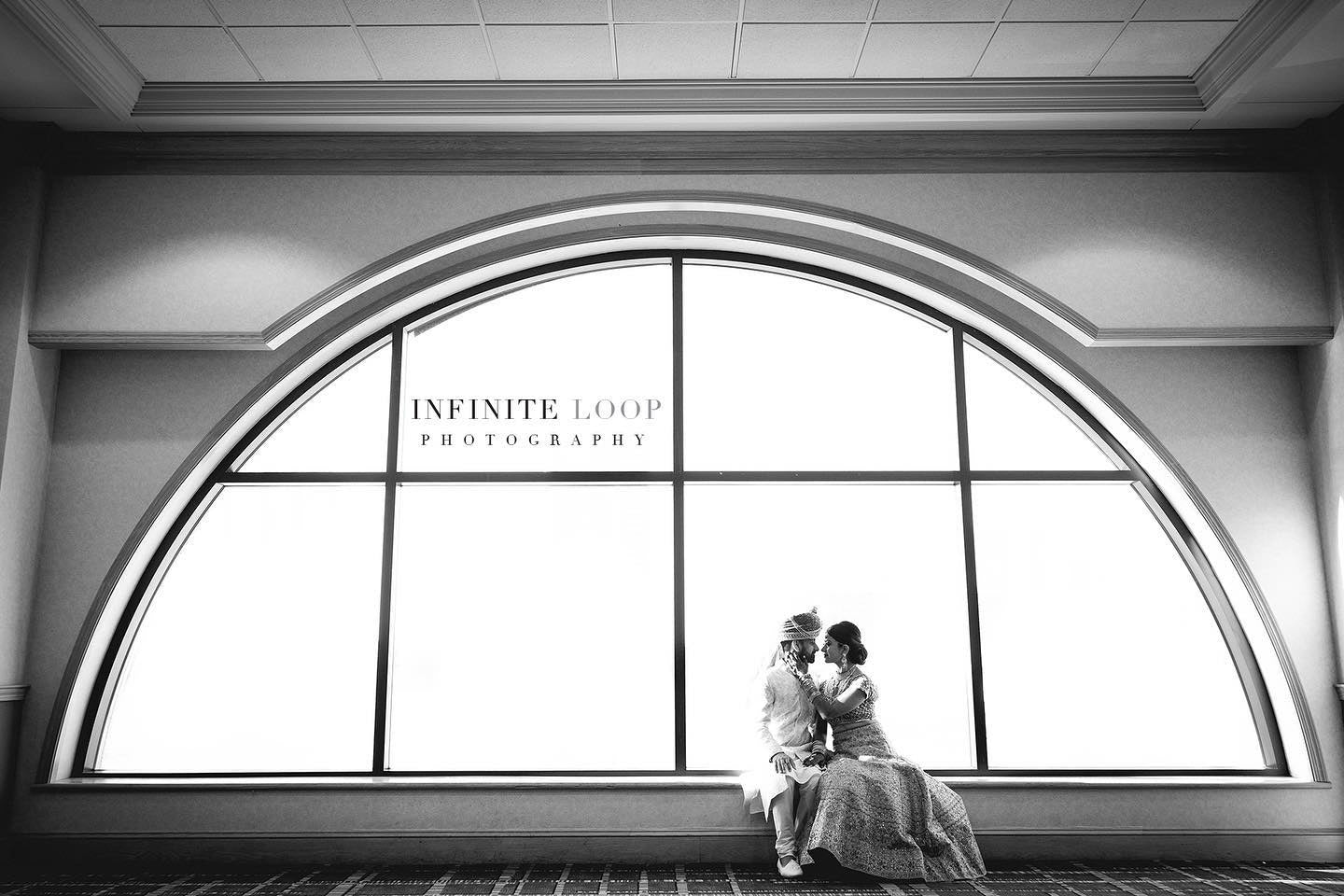 a wedding couple sitting in their wedding attire in front of a big arching window