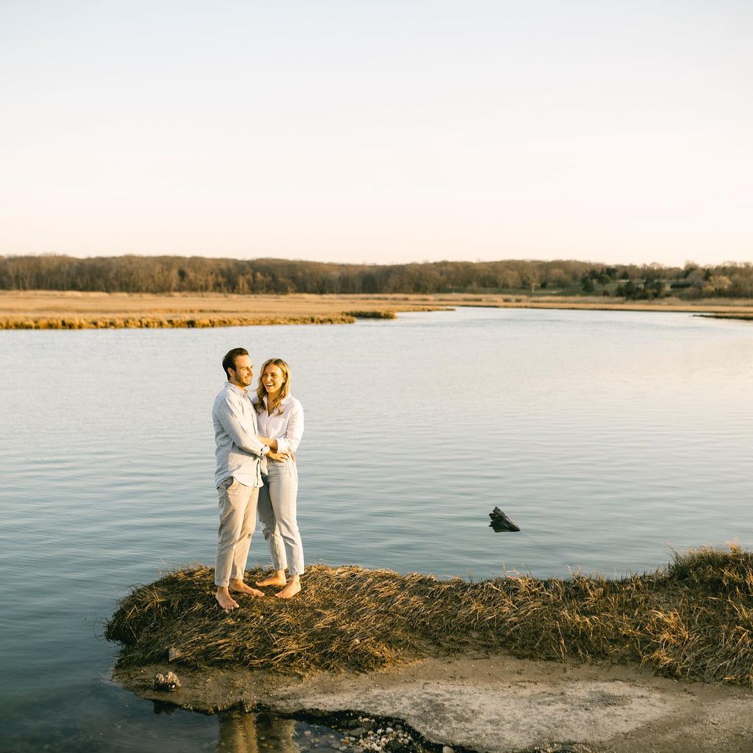 an engaged couple standing by a lake holding each other