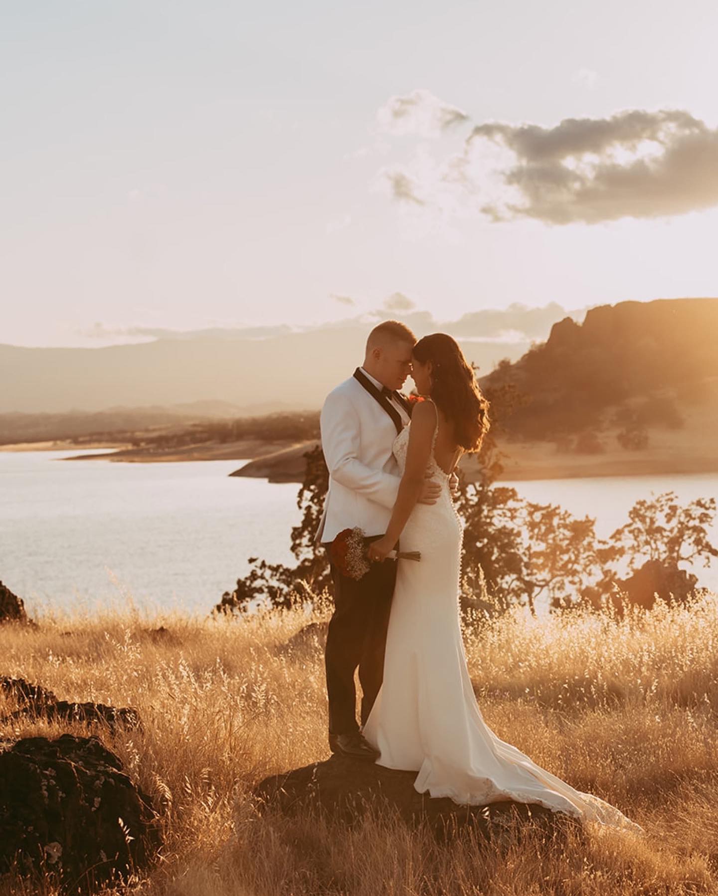 a couple standing on a grassy terrain overlooking a lake during sunrise