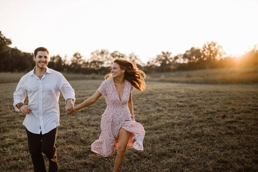 a couple running through a meadow holding hands while the sun is setting behind them