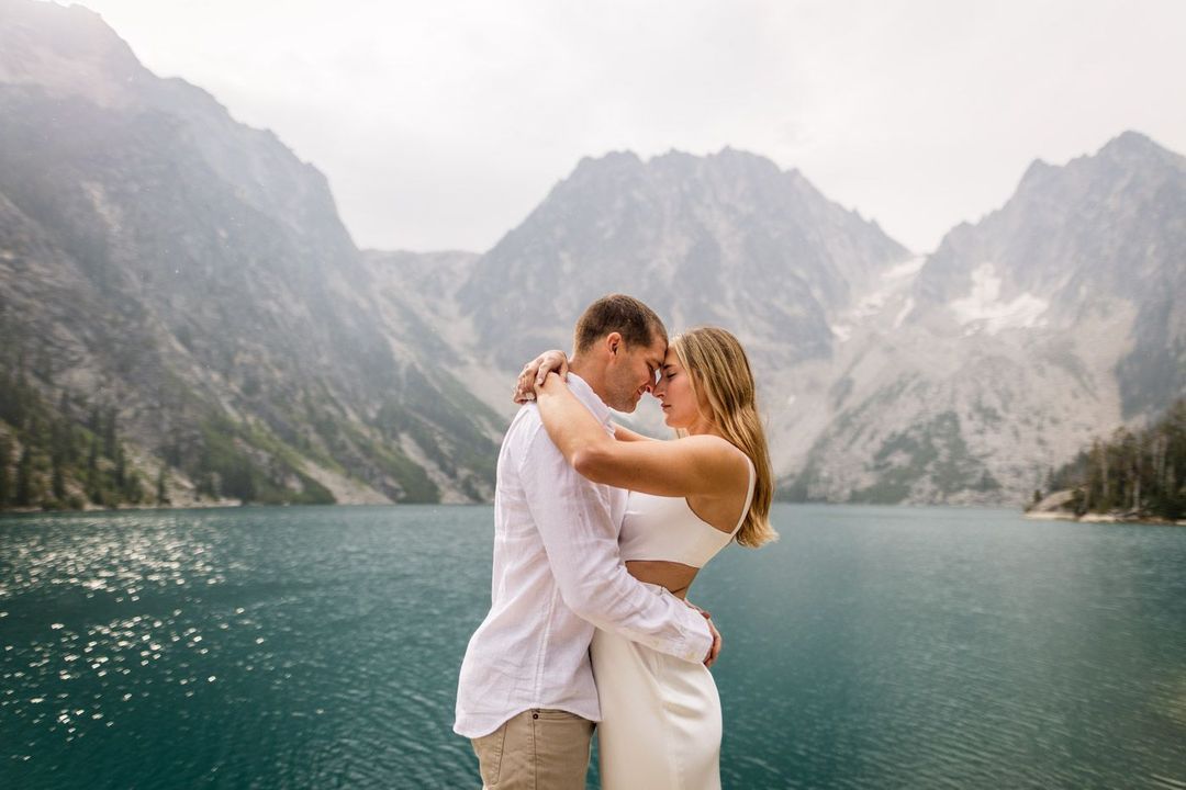 a couple touching their heads together standing by a lake surrounded by mountains
