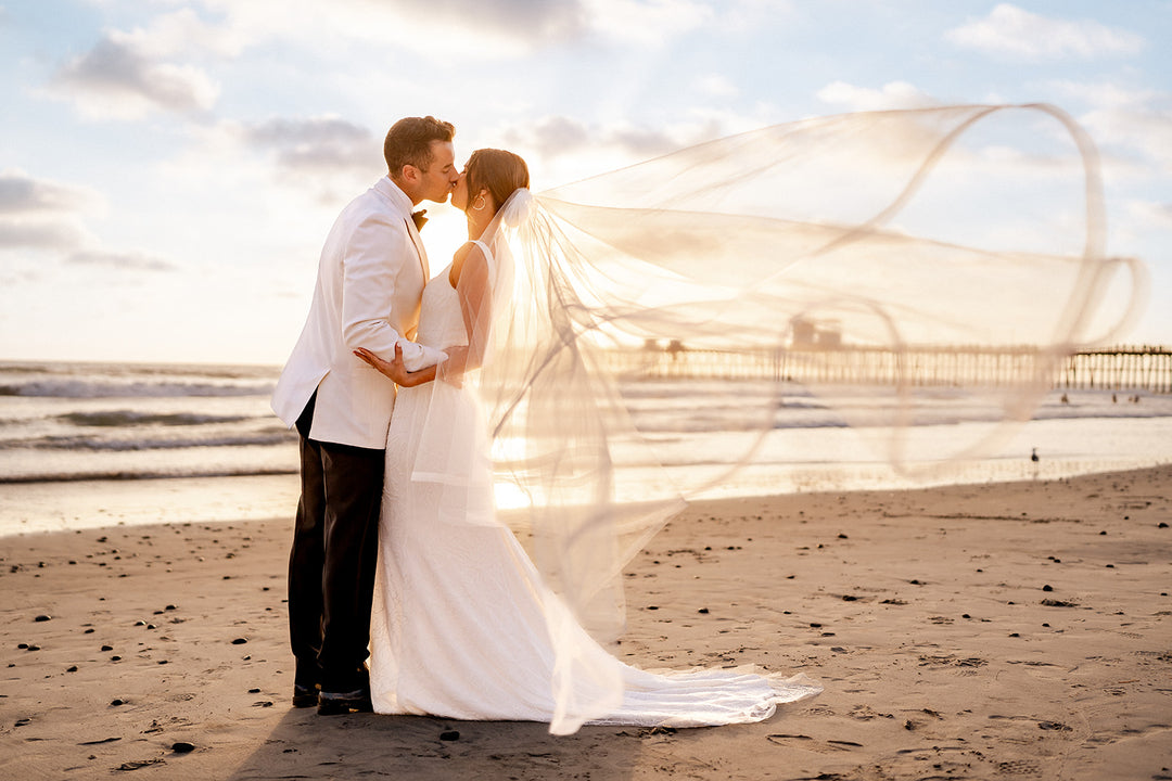 a newlywed couple kissing on the beach while the brides wedding veil blows behind her