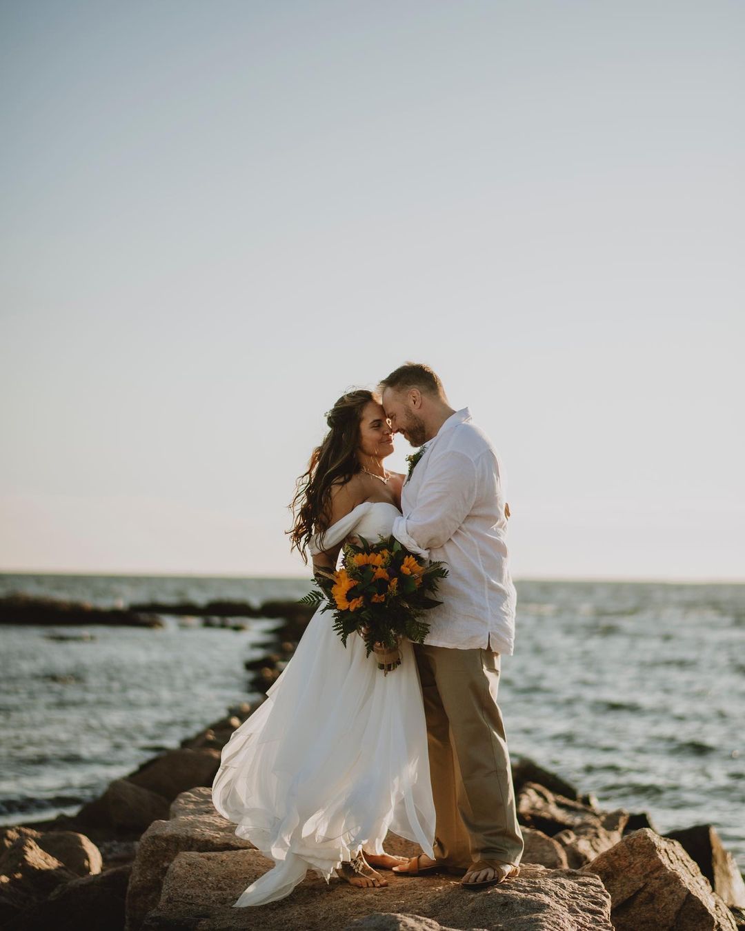 a wedding couple standing on a small pile of rocks beside the sea