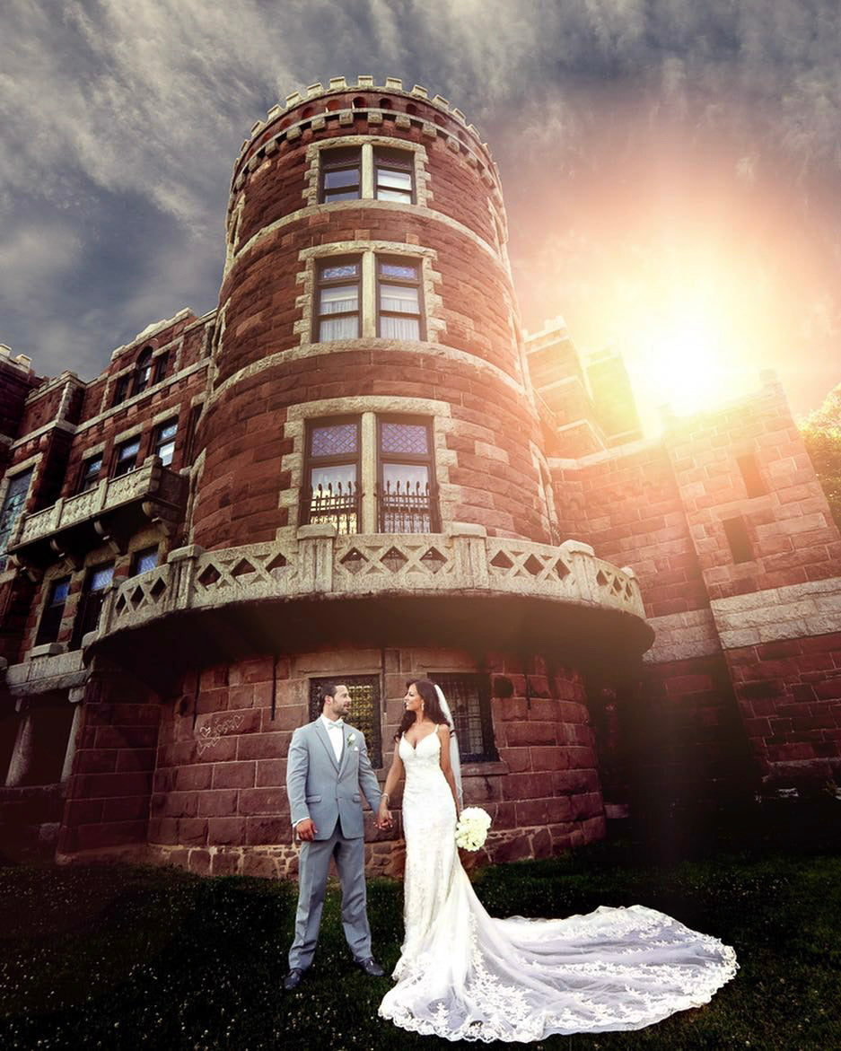 a wedding couple standing holding hands in their wedding attire while the sun is shining from behind a building