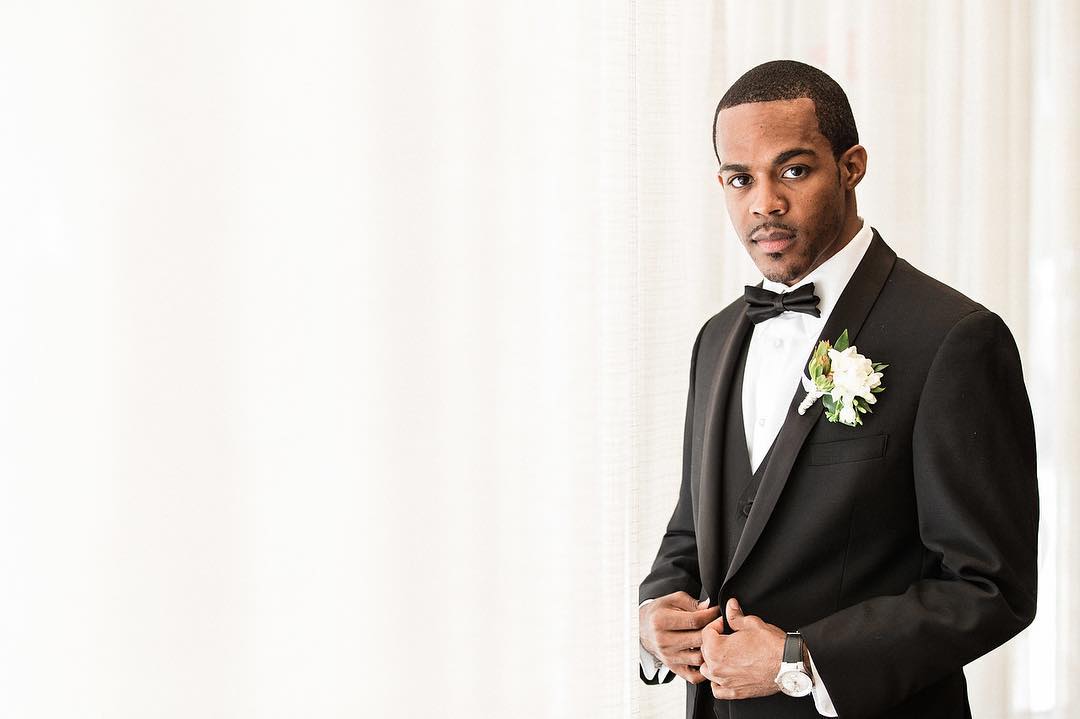 A groom posing for a picture in a black tuxedo with white flower boutonnieres