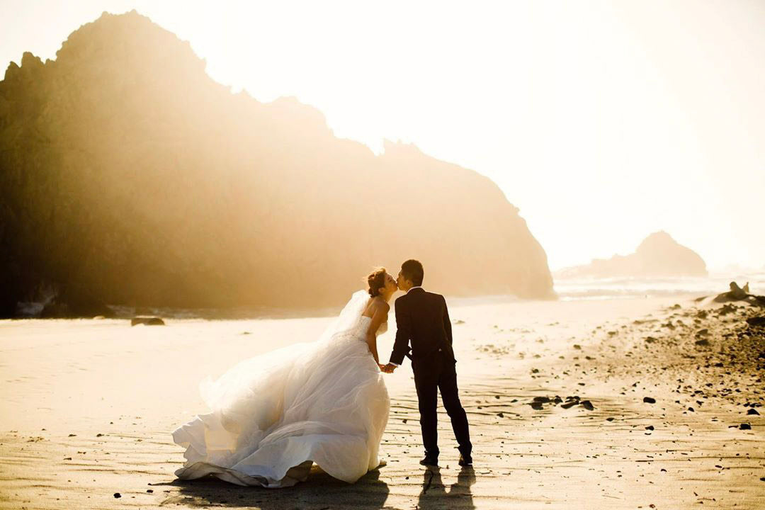 a wedding couple kissing by a beach while the sun is setting behind them