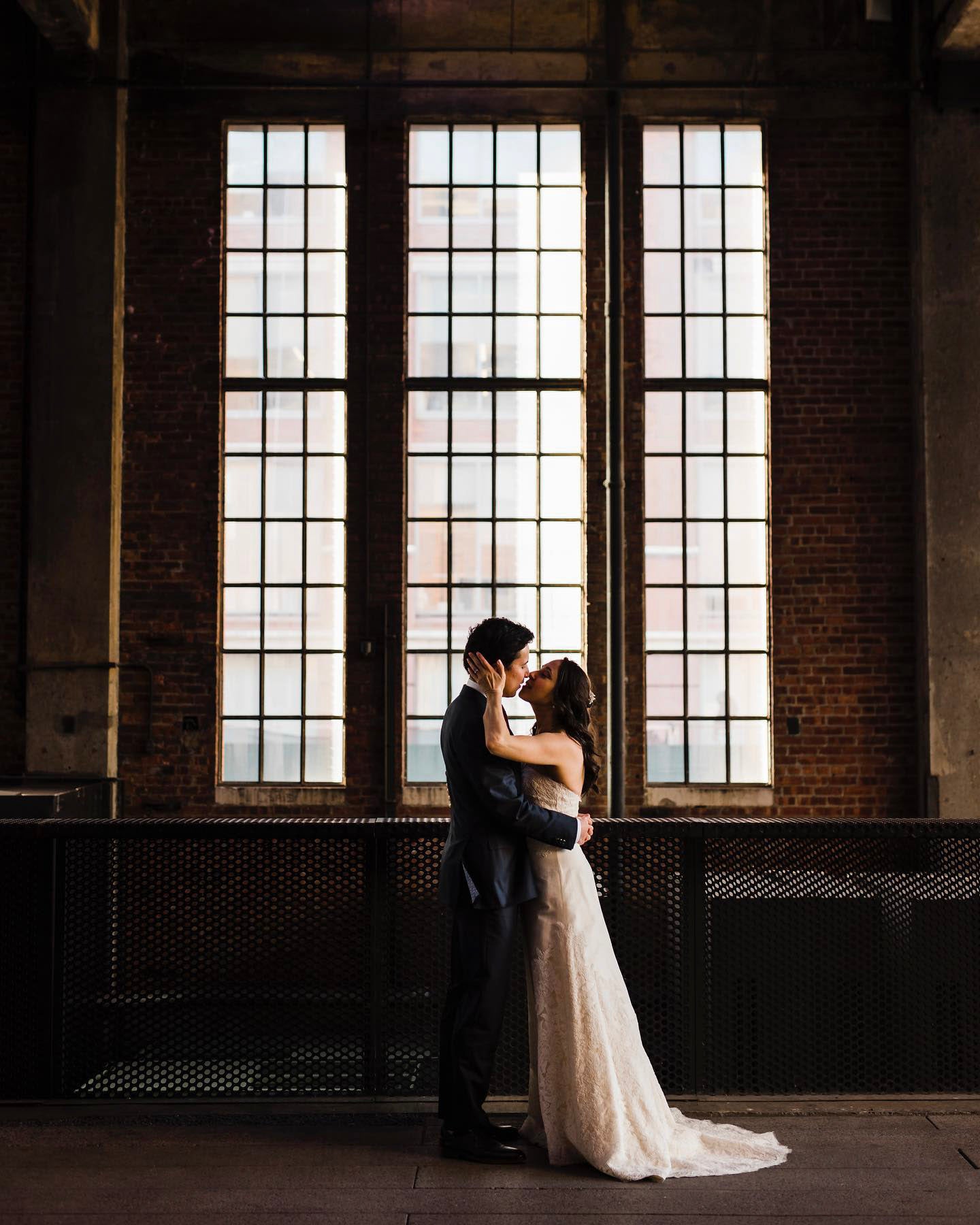 a newly wed couple kissing in front of a large window in a low light setting
