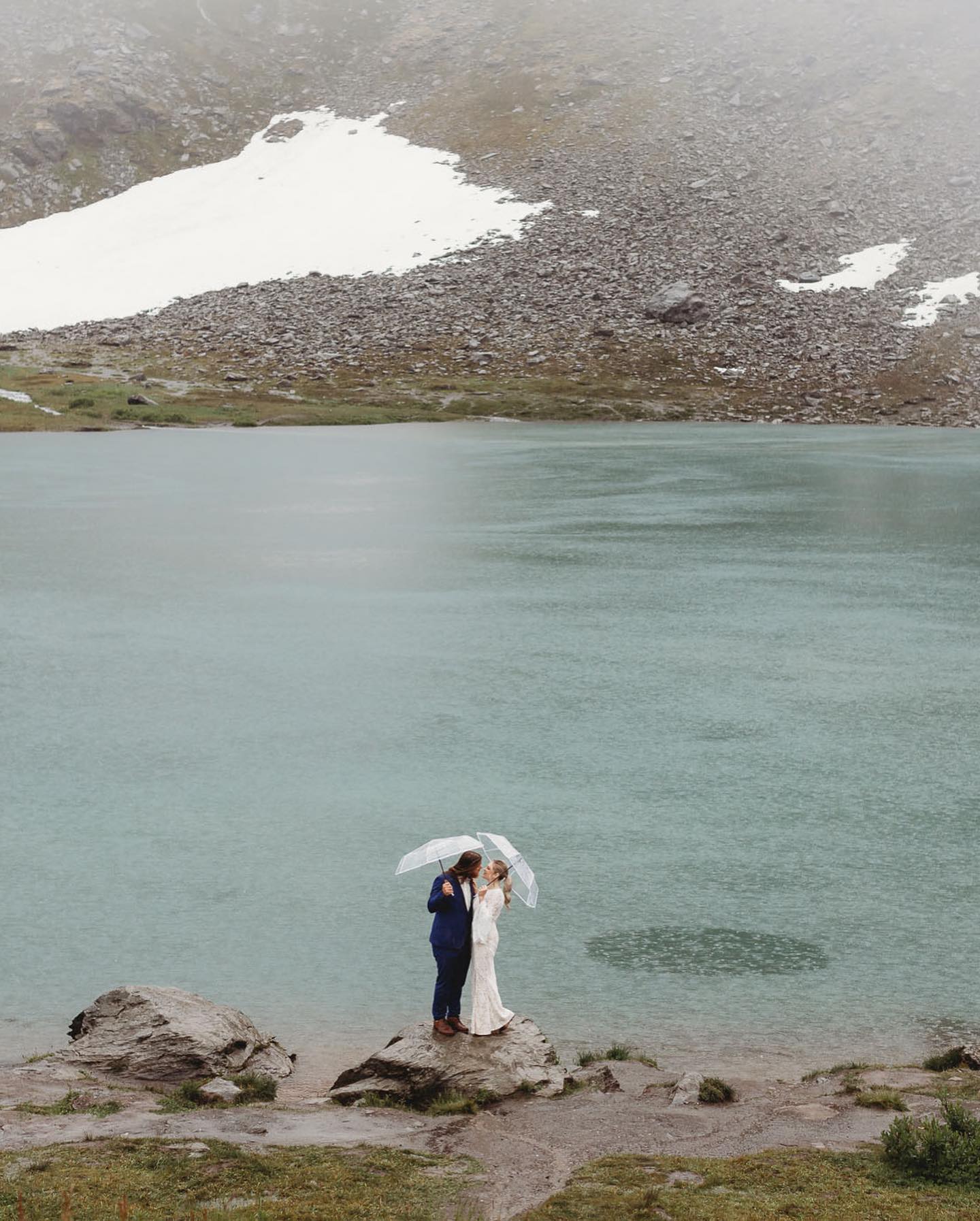 a couple standing by a lake holding umbrellas in their wedding attire while its raining