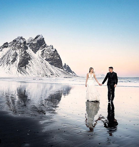 a wedding couple holding hands and walking on a beach in front of a snow capped hill