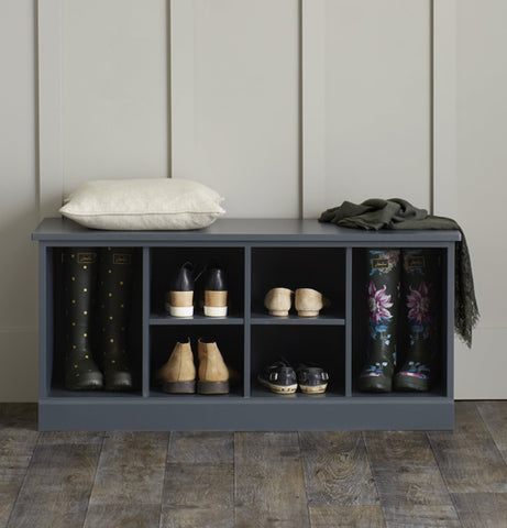A dark blue shoe bench against a grey, wood panelled wall. On top of it sits an off-white coloured cushion and a wool scarf.