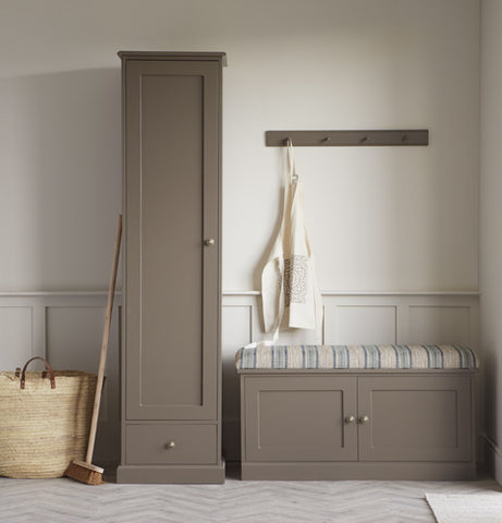 A tall, grey-brown utility cupboard stands next to a two-door shoe bench of the same colour. To the left is a wooden sweeping brush and woven wicker basket leaning against the wall.