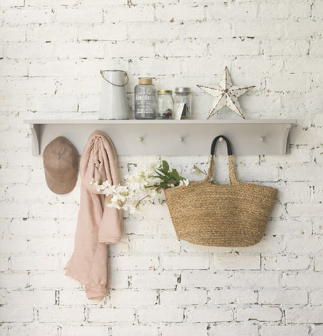 A light grey, wooden peg rail displays a wicker tote bag, pink scarf, brown baseball cap and various glass jars, against the backdrop of a distressed, white, brick wall.