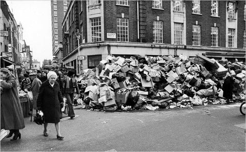 A black and white photograph shows an elderly woman walking across a road in front of piles of rubbish, lining the road during the garbage collectors strike in London during the 1970s