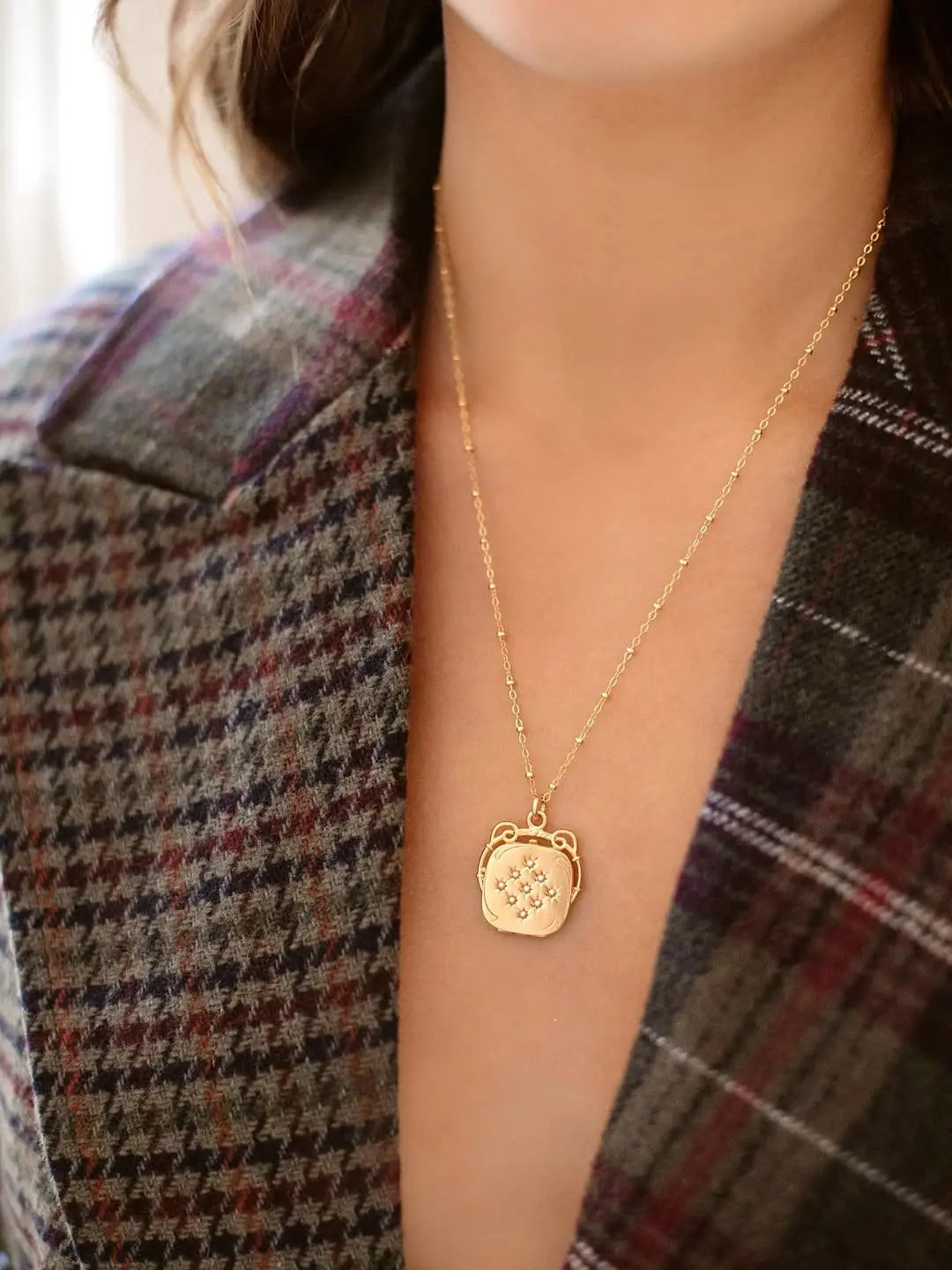 Woman wears a grey and purple plaid blazer and a gold chain necklace with an ornate square locket featuring 9 small gems.