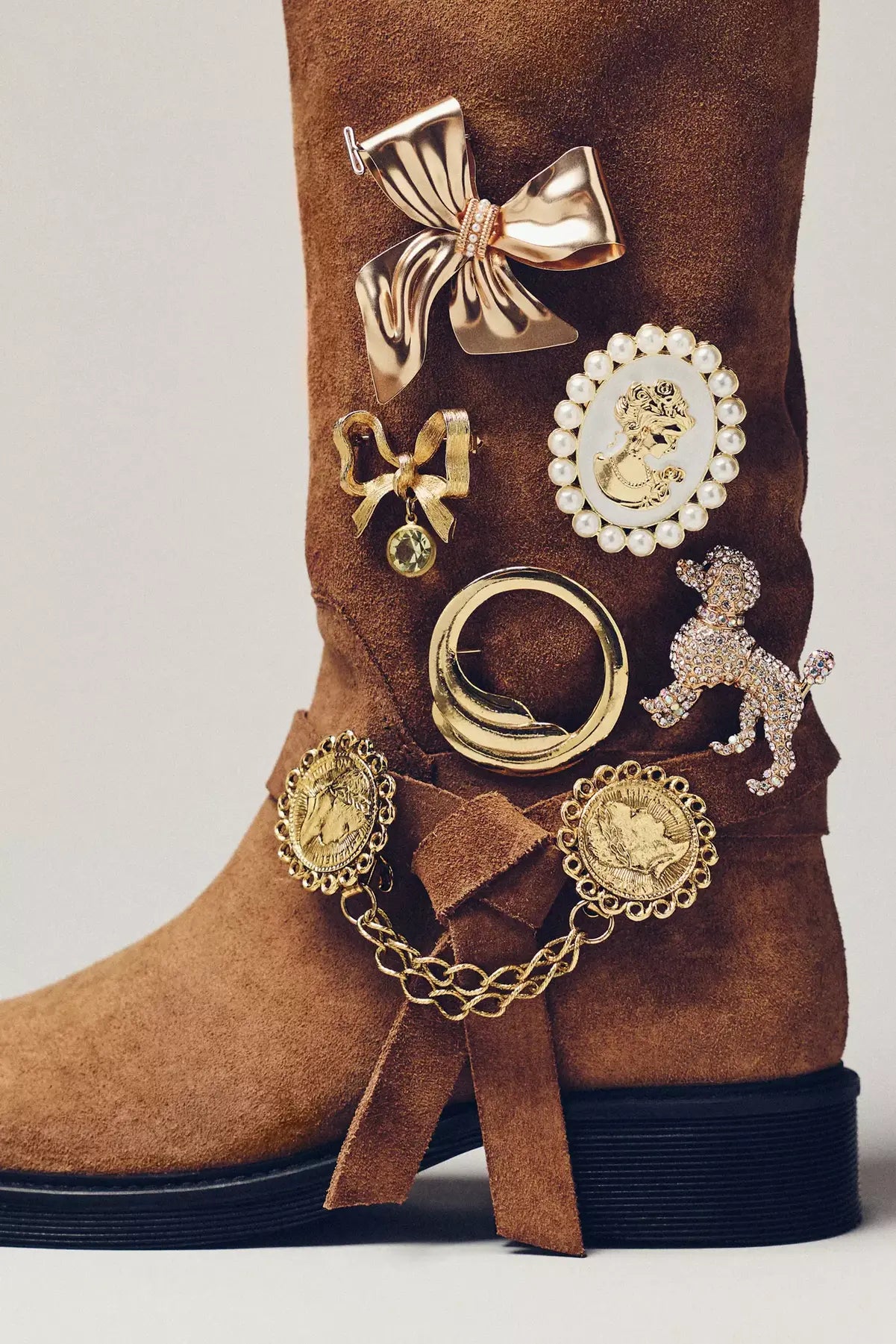 Brown suede boot adorned with various gold-tone brooches: large bows, a pearl-edged cameo, a crystal poodle, an abstract ring, and two coin pendants.