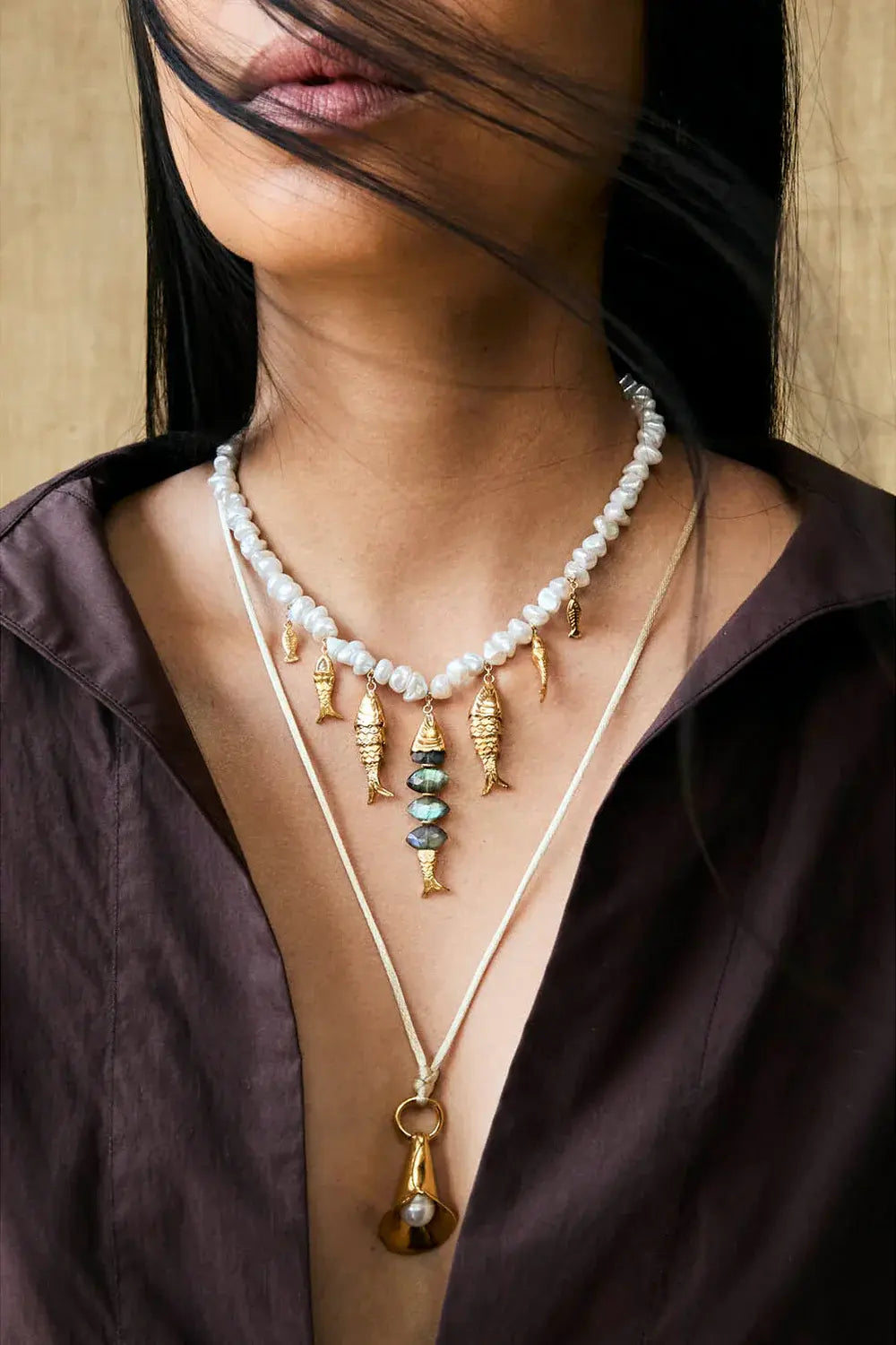 Woman in a brown shirt wears a pearl necklace with gold fish and green stone charms, and a longer necklace with a gold bell pearl pendant.