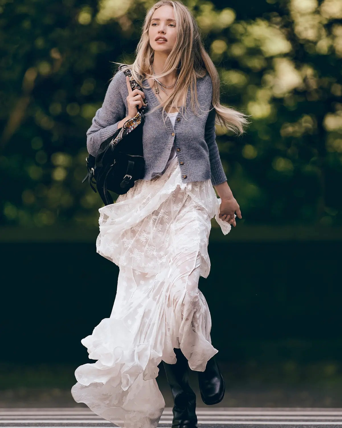 Woman in a gray knit cardigan, white camisole, flowing white lace skirt, and black boots, holding a black shoulder bag, outdoors.