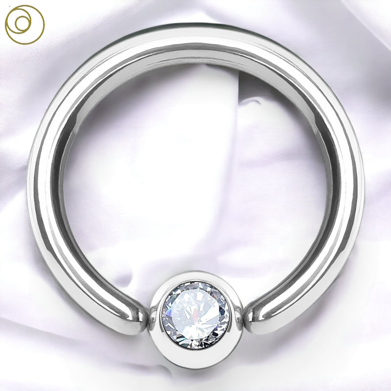 A 16g conch piercing ring with a fixed ball that has a clear gem on one side of the fixed ball. It is pictured on against a white fabric background.