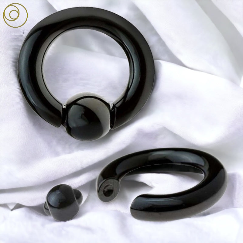 Two views of a black captive bead ring. One captive ring has the spring action ball removed and the other has it in. Both are pictured against a white fabric background.