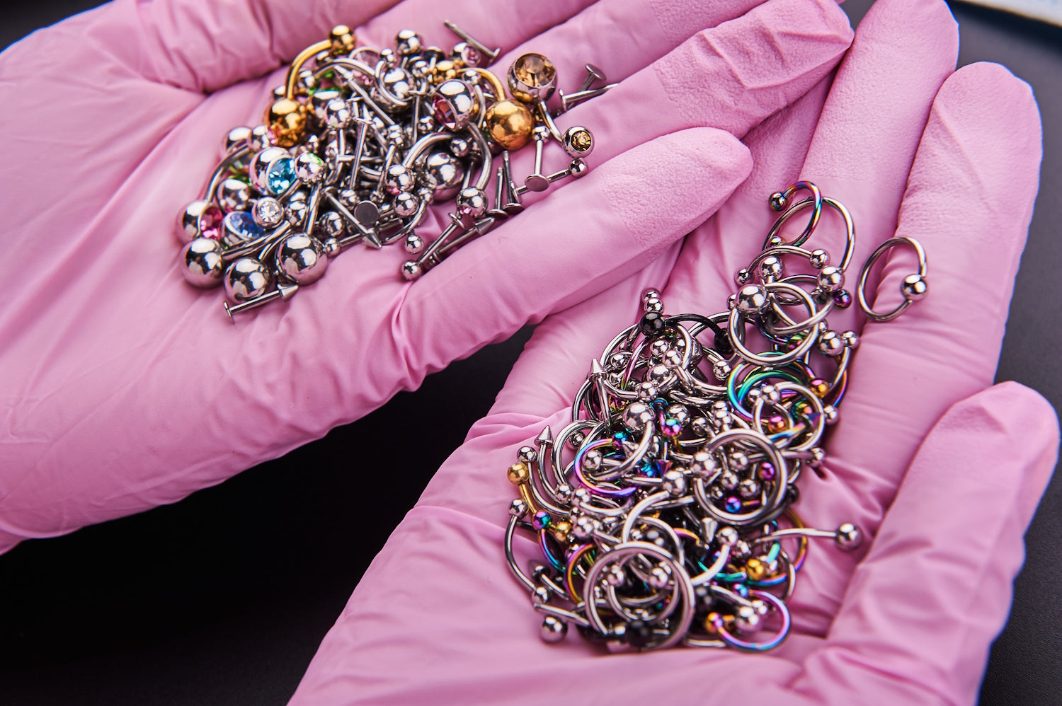 A person with pink gloves on that has many pieces of body piercing jewelry in her hands including titanium and gold pieces.