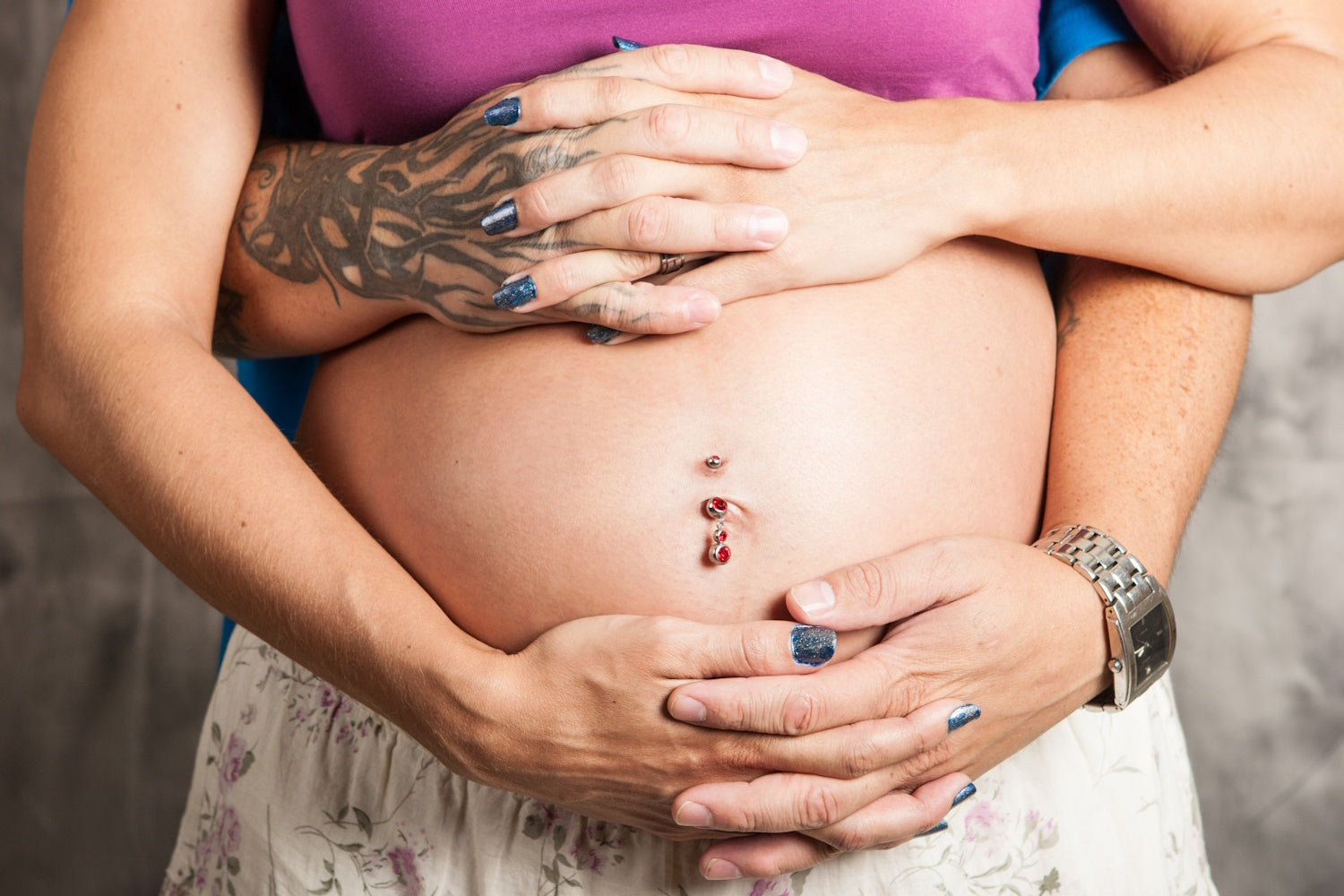 A pregnant woman with a belly button piercing with a persons arm around her.