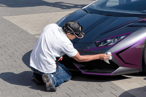 An individual wearing a cap and gloves cleans the front of a luxurious purple vehicle on a paved surface.