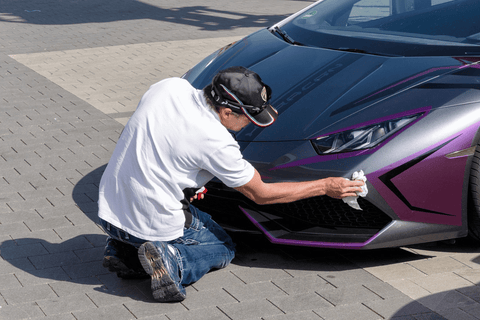 A person in a white shirt and jeans kneeling while polishing the grille of a purple sports car.