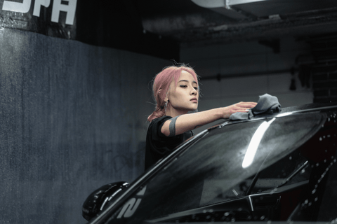 A person with pink hair and a black top reaches over the hood of a car, polishing it with a gray cloth.
