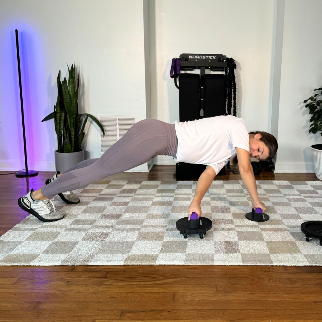 Person doing a plank exercise with fitness equipment on a checkered rug indoors.