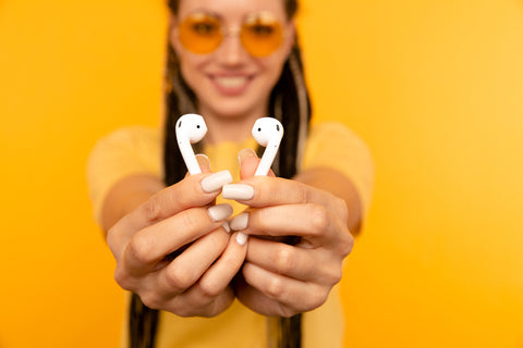 Woman in sunglasses holding pair of white Apple Airpods, standing in front of yellow background.