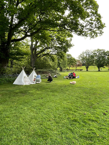 A wide shot of gardens with big trees and bushes and a big grassed area. Two children’s play teepees sit on the grass with a group of people sitting like a picnic