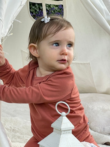 Toddler in merino wool pyjamas sits holding the side of a teepee and looks up. There is a candle holder in front.