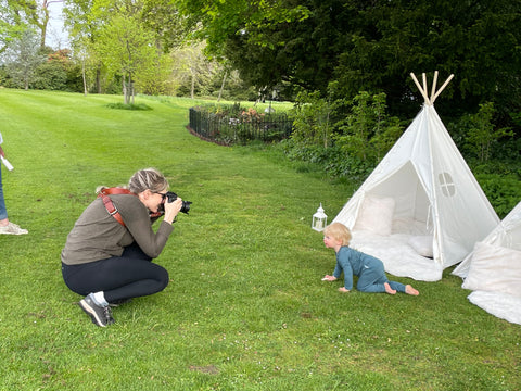Open lawn with a woman crouched taking a photo of a toddler crawling towards her. Play teepees and big garden in the background.
